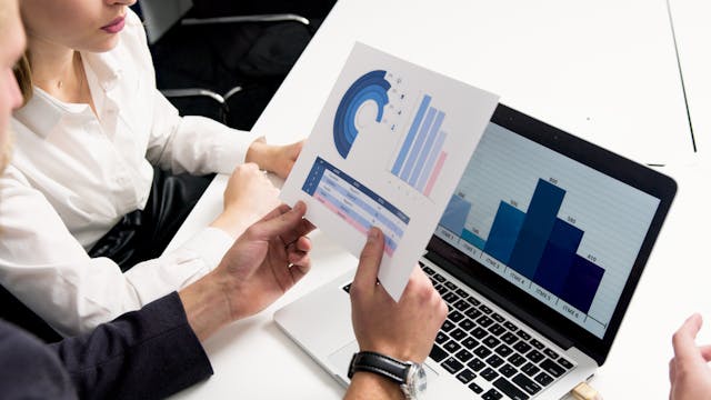 Colleagues discuss printed analytics beside a laptop displaying a bar graph.