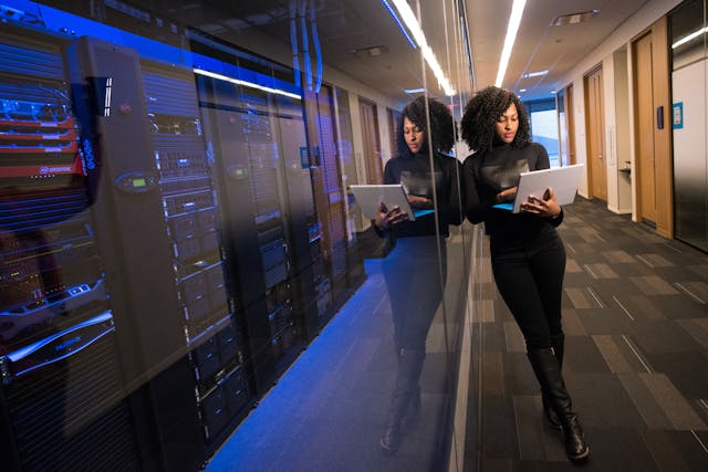 IT engineer leans against a server-room glass wall while working on a laptop.