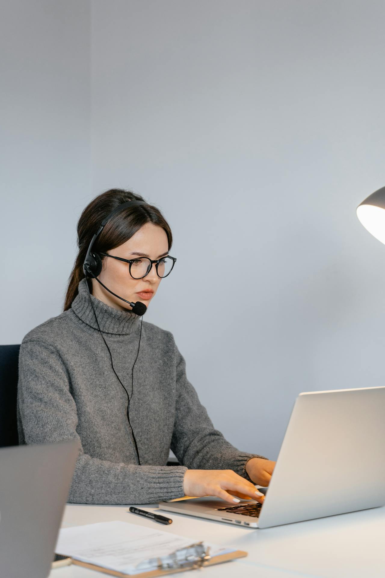 Woman in glasses and headset types on a laptop at a minimalist workstation.