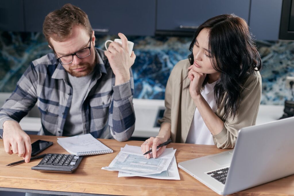 Worried couple use a calculator and laptop to work through bills at their table.