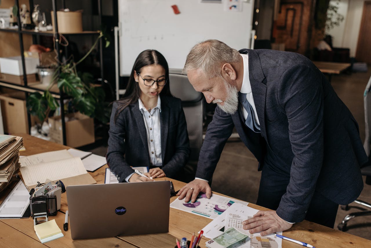 Desk-side review – In a modern office, a seated woman and an older man in suits examine printed charts spread across a desk with an open laptop.