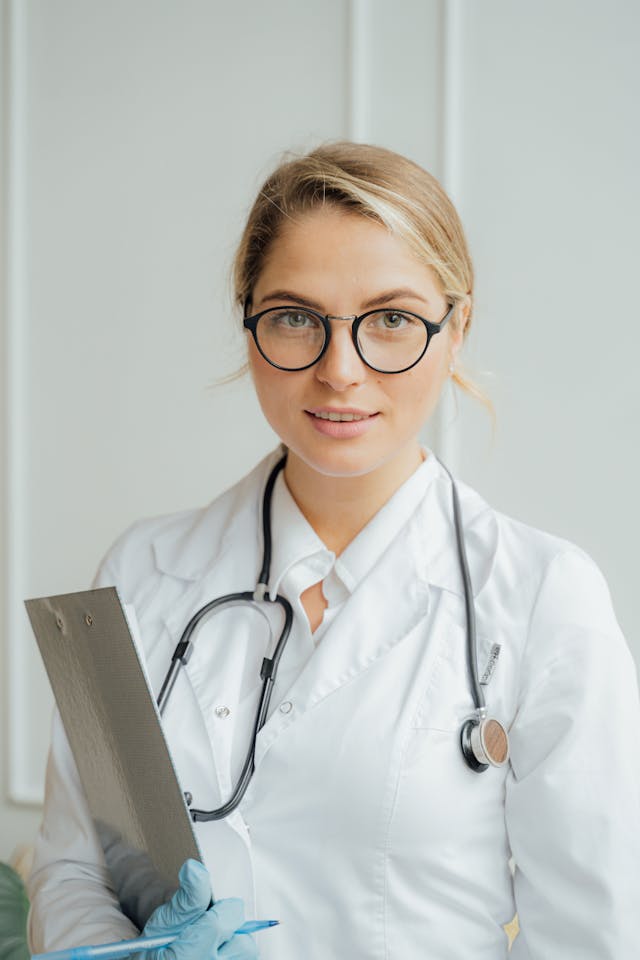 Healthcare professional – A woman in a white lab coat with a stethoscope and clipboard, wearing round glasses and blue examination gloves.