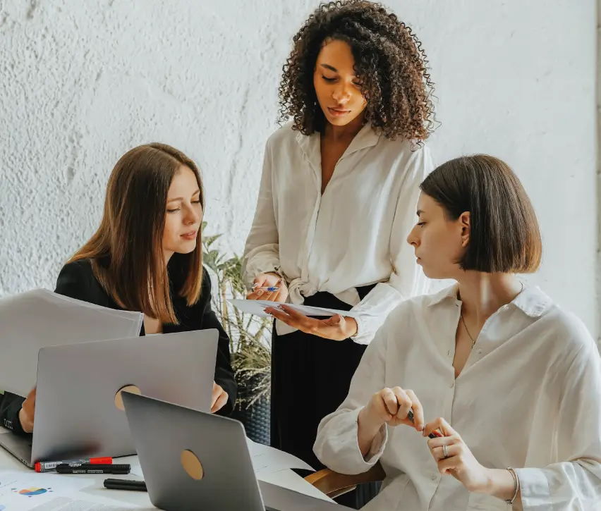 Small group collaboration – Three women in business-casual outfits gather around laptops and documents; one is standing and pointing something out on a tablet while the others listen.